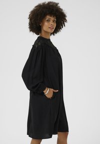 Robe noire à manches longues avec un col haut et des détails en dentelle sur les épaules. Dispose de manches larges, de poches et d'une coupe décontractée.