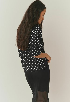 Sort bluse med hvide prikker, trekvartærmer, afslappet pasform og sideslidser, båret over et sort skørt med transparente paneldetaljer.