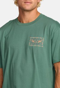 Groene katoenen T-shirt met korte mouwen, gekenmerkt door een bloemenafdruk patch graphic in warme kleuren op de linkerborst. Zachte textuur.