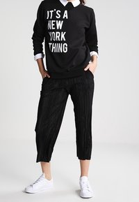 Sudadera negra con texto blanco, combinada con pantalones recortados negros con plisados. El conjunto se completa con zapatillas blancas.