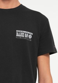 Černé tričko s krátkým rukávem a bílým nápisem "Tommy Jeans Bank of NYC 1985" potisknutým na levém prsu.