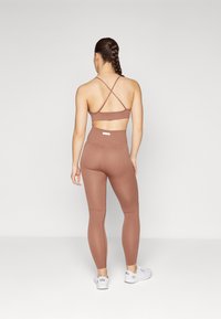 Gestreifter, brauner Sport-BH mit Cross-Back-Design, kombiniert mit hochgeschnittenen, gestreiften braunen Leggings. Modell trägt weiße Sneakers.
