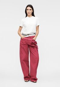 Γυναίκα που φοράει λευκό κοντομάνικο πουκάμισο και κόκκινο παντελόνι με φαρδιά πόδια και κεντήματα με ανθικά μοτίβα, στέκεται με τα χέρια στις τσέπες σε γκρι φόντο.