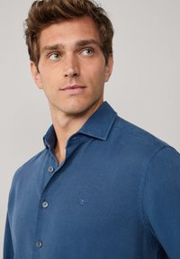 Giovane uomo con capelli castano chiaro che indossa una camicia azzurra con bottoni e un piccolo logo ricamato sul petto, che guarda leggermente di lato.