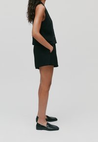 Czarny bezrękawnik z okrągłym dekoltem i kieszonką, zestawiony z czarnymi szortami o surowym wykończeniu. Całość uzupełniają czarne loafery.