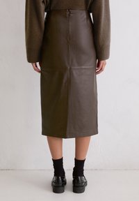 Brauner Ledermidirock mit Naht-Design, der eine glatte Textur und einen diskreten Reißverschluss hinten aufweist. Kombiniert mit schwarzen Socken und Schuhen.