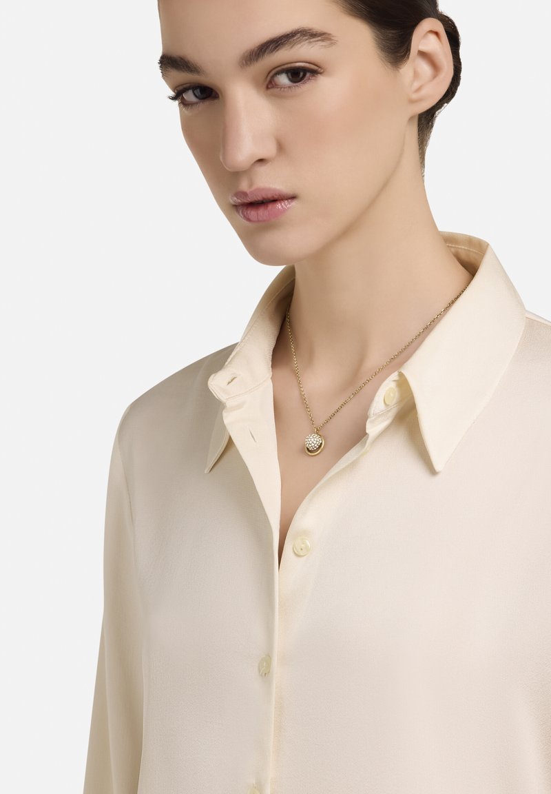 Blouse crème avec un col classique, une texture douce et des boutons sur le devant. Collier en or avec un pendentif circulaire texturé.