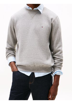 Man in een lichtgrijze sweatshirt over een blauw gestreepte shirt met donkere jeans, hand in de zak, tegen een witte achtergrond.