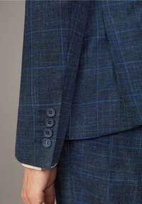 Gros plan sur le bras d'un homme portant une veste de costume à carreaux bleu foncé avec quatre boutons au poignet et un pantalon assorti.