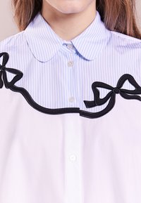Biała koszula z jasnoniebieskim i białym paskowaniem kołnierzyka, z czarnymi akcentami w postaci kokard oraz wykończeniem wzdłuż dekoltu. Z przodu widoczne guziki.