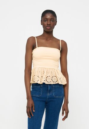 Γυναίκα σε όρθια στάση, φορώντας μπεζ αμάνικο τοπ με διακοσμητική λεπτομέρεια στο τελείωμα και μπλε τζιν, σε απλό λευκό φόντο.