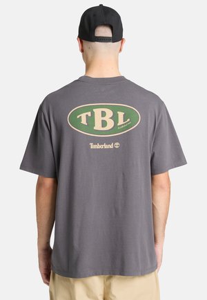 Szary bawełniany t-shirt z dużym zielono-beżowym owalnym logo z napisem „TBL” i „Timberland” na plecach. Krótkie rękawy, luźny krój.