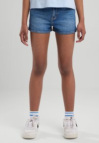 Pantaloni shorts in denim di un blu medio, design aderente, orlo sfilacciato, abbinati a sneakers bianche e calze a righe blu.