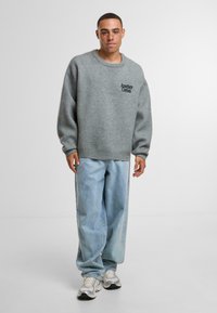 Maglione grigio lavorato a maglia con scollatura rotonda e piccolo logo "Another Cotton". Abbinato a jeans baggy azzurri e scarpe da ginnastica comode.