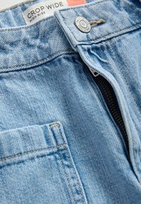 Világoskék magas derekú, széles szárú farmernadrág részben nyitott cipzárral, fém gombbal és elől zsebrátétes díszítéssel.