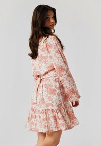 Robe à motifs floraux en rose clair et blanc, avec des manches longues, une taille cintrée et un ourlet festonné. Une ceinture nouée accentue la silhouette.