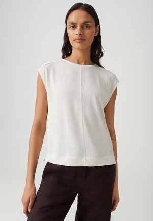 Femme aux longs cheveux foncés portant un haut blanc sans manches et un pantalon foncé, debout devant un fond clair uni.