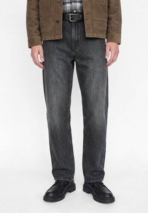 Vue recadrée d’un mannequin debout portant un jean gris foncé, une ceinture noire, une veste marron, une chemise à carreaux et des chaussures noires.