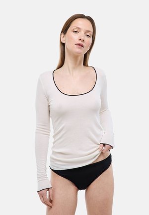 Femme aux cheveux bruns raides portant un haut blanc à manches longues avec bordure noire et une culotte noire, posant devant un fond blanc.