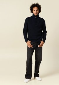 Pull en maille bleu marine avec un col montant et une fermeture éclair quart, associé à un jean noir et des baskets blanches, sur un fond neutre.