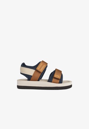 Navy and tan sandals with adjustable straps, textured fabric, soft sole, and a contrasting white midsole. Suitable for warm weather.