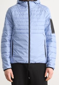 Ljusblå pufferjacka med huva och diagonal quiltad design. Har svart dragkedja, sidofickor och en logotypapplikation på ärmen.