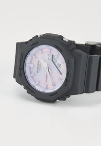 Montre G-Shock noire avec un boîtier rond texturé, un cadran rose clair, des index horaires en relief et un affichage numérique. Bracelet en caoutchouc avec boucle en métal réglable.
