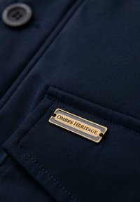 Detail van een marineblauwe stoffen jas met een metallic gouden label dat "OMBRE HERITAGE" zegt, met een enkele zwarte knoop boven de zak.