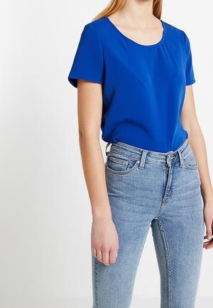 Kobieta ubrana w jasnoniebieską bluzkę z krótkim rękawem wpuszczoną w jasnoniebieskie, wysokowalne dżinsy na tle jednolitego białego tła.