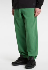 Groene sweatpants gemaakt van zacht materiaal, met een relaxte pasvorm, elastische boorden en een gladde textuur, gecombineerd met zwarte sneakers.