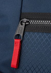 Pogled na mornarsku tkaninu koja prikazuje zatvarač s crnim zubima, crni najlon odjeljak i crvenu traku za povlačenje s brandiranjem "TOMMY JEANS".