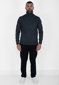 Maglione a collo alto grigio scuro con polsini e orlo a coste, abbinato a pantaloni neri e sneakers azzurro chiaro, su uno sfondo bianco.