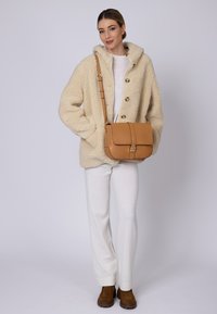 Beige fleece jakke med knapper, cremefarvet sweater, hvide bukser og brune støvler. Den tanfarvede crossover-taske har en klap og metalspænde.