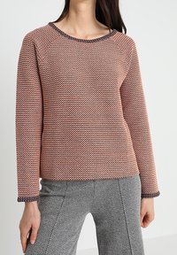 Sweter koralowy na drutach z teksturowanym wzorem, okrągłym dekoltem oraz kontrastującymi ciemnymi wykończeniami na mankietach i dekolcie, zestawiony ze spodniami w jodełkę.