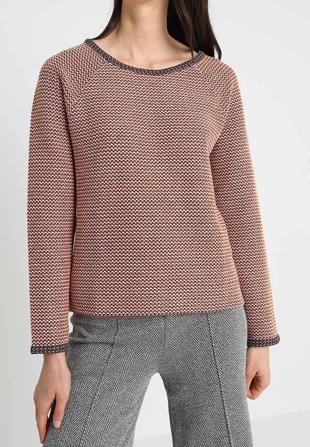 Sweter koralowy na drutach z teksturowanym wzorem, okrągłym dekoltem oraz kontrastującymi ciemnymi wykończeniami na mankietach i dekolcie, zestawiony ze spodniami w jodełkę.