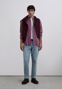 Dunkelrotes Hemd mit Knöpfen, darüber ein dunkler Pullover, hellblaue Jeans und braune Stiefeletten. Das Model steht vor einem neutralen Hintergrund.