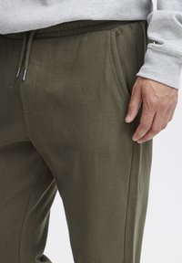 Olivgrüne Jogginghosen aus einem weichen Material, mit elastischem Bund und Kordelzug sowie seitlichen Taschen; getragen mit einem grauen Langarmshirt.