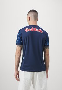 Maglietta da atletica blu navy a maniche corte, con un grande logo rosso "Red Bulls" sulla schiena e dettagli blu sulle maniche. Texture liscia.