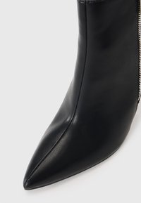 Bottine en cuir noir à bout pointu avec couture avant visible et fermeture éclair latérale argentée sur fond neutre.