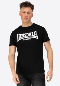 Melnas kokvilnas t-krekls ar lieliem baltiem "LONSDALE LONDON" logotipiem treknā, izliekta tekstā, kas izvietots visā krūtīs. Īsas piedurknes un apaļa kakla izgriezums.