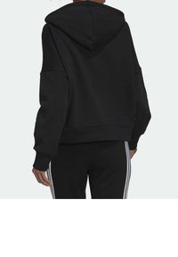 Sweat-shirt à capuche noir avec épaules tombantes et poignets côtelés. Tissu doux, coupe courte. Porté avec un pantalon de jogging noir assorti avec des bandes blanches.