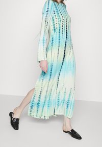 Dlouhé, volné, batikované šaty v modrých a zelených tónech s vertikálními vzory; spojené s černými, strukturovanými klouzavými mules.