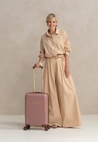Een lachende vrouw in een beige outfit staat naast een roze koffer met "SUITSUIT" erop geschreven, tegen een neutrale achtergrond.