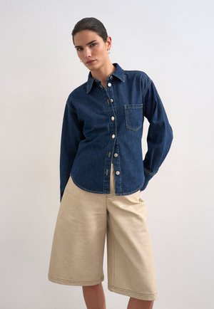 Γυναίκα που φορά σκούρο μπλε τζιν πουκάμισο με κουμπιά και μπεζ φαρδιά σορτς μέχρι το γόνατο, στέκεται μπροστά σε απλό φωτεινό φόντο.