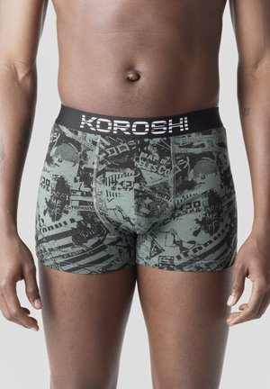 Torso masculino vestido con bóxers grises con patrones abstractos negros y texto "KOROSHI" en la pretina, sobre un fondo liso.