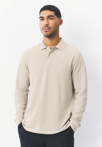 Pitkähihainen beige poolopaita, valmistettu pehmeästä kankaasta, jossa on klassinen kaulus ja hillitty rintalogo. Yhdistetty tummiin housuihin.