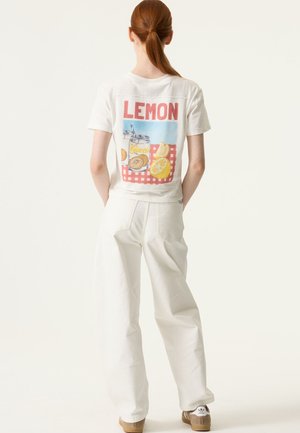 Camiseta blanca de algodón con un gráfico colorido de limón en la espalda, sobre un fondo de cuadros rojos; combinada con pantalones blancos de tiro alto.