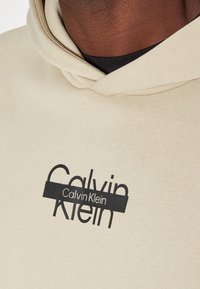Felpa beige con una morbida texture, caratterizzata da un logo nero ben visibile con la scritta "Calvin Klein" sul petto e un cappuccio con coulisse.