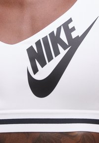 Κοντινή λήψη κορμού που φοράει λευκό αθλητικό σουτιέν Nike με μεγάλο μαύρο λογότυπο Nike και το σκούρ στο στήθος.