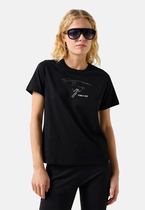 Blonde vrouw met grote zwarte zonnebril en een zwart T-shirt met een skiër en de tekst "FIRE+ICE" op de voorkant.